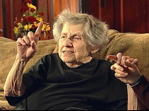 The image is from a video, Worry,  about a DB woman who survived the Holocaust. It shows a DB woman with grey short hair wearing a black top signing and sitting on a beige sofa.